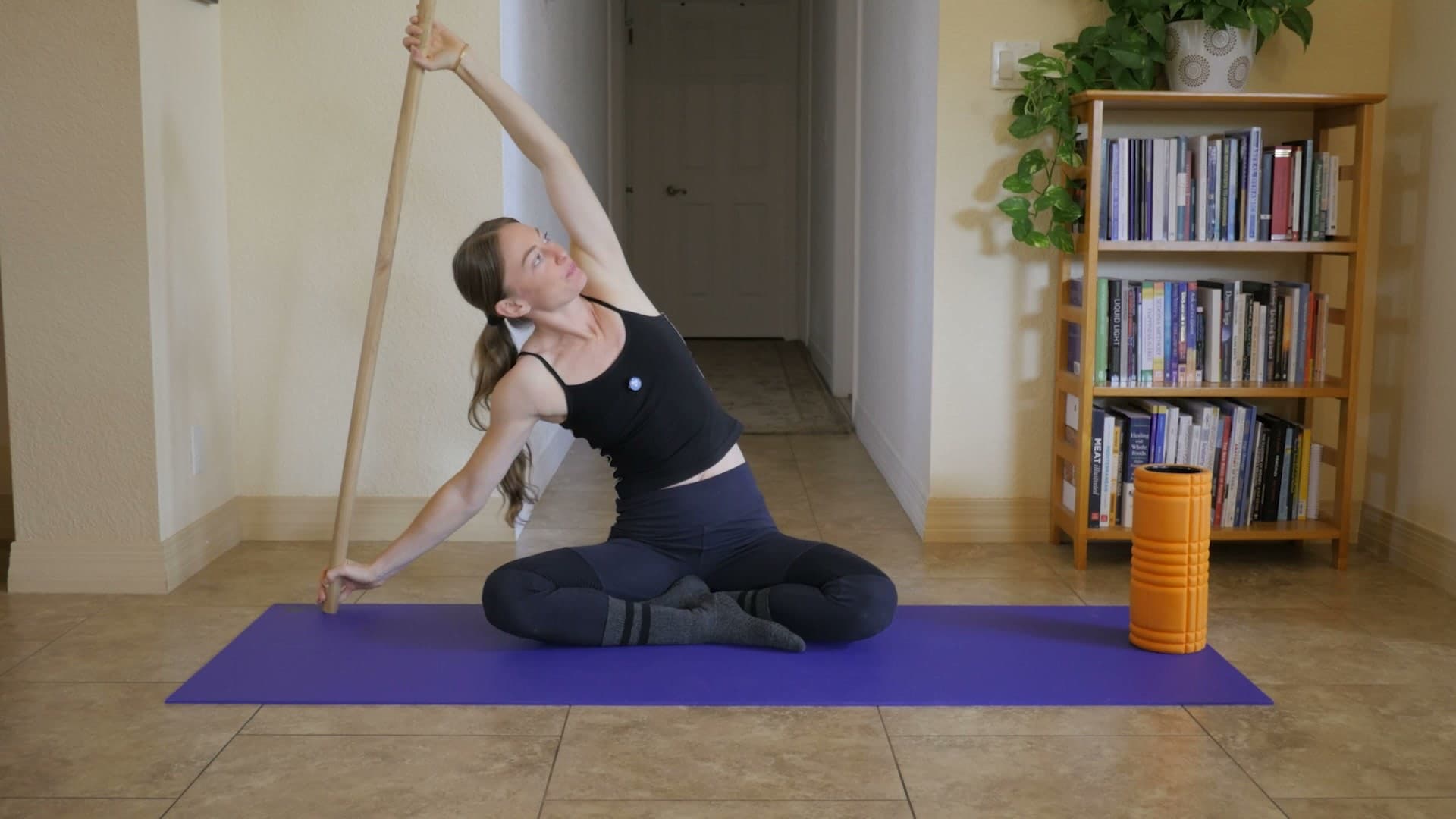 Seated cross-legged on a mat, bending to the side with a dowel held overhead